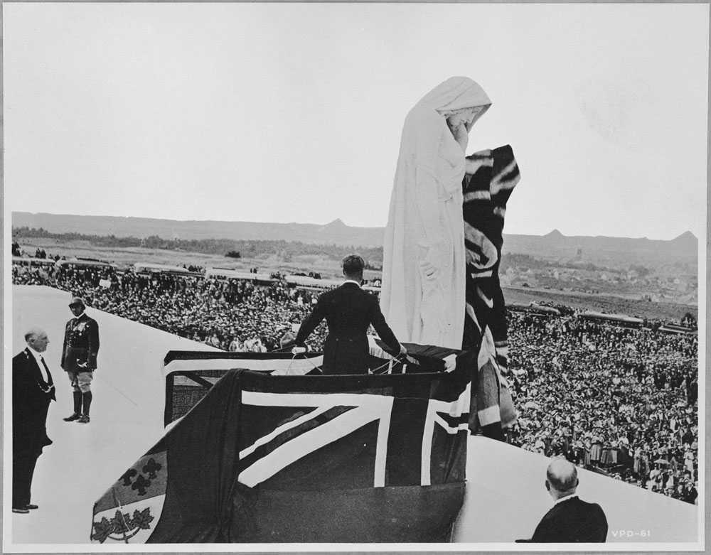 Black and white photograph. King Edward III, back to the camera, wearing a tuxedo, uses ropes to unveil the  Mother Canada figure, covered in the Union Jack and Red Ensign.  A large crowd looks on.